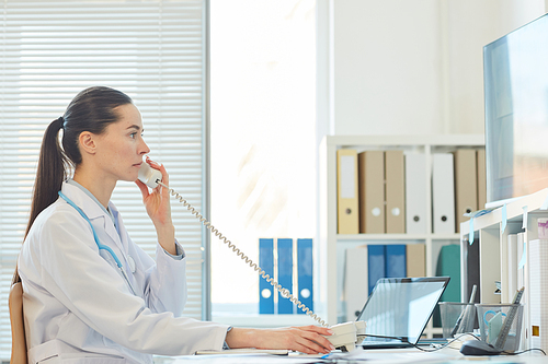 Side view portrait of serious female doctor speaking by phone while sitting at desk and working in clinic or hospital, copy space