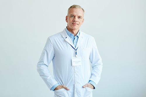 Waist up portrait of confident mature doctor wearing lab coat and  while standing against white wall in clinic, copy space
