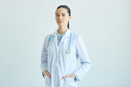 Waist up portrait of confident female doctor wearing lab coat and  while standing against white wall in clinic, copy space