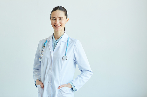 Waist up portrait of friendly female doctor wearing lab coat and  while standing against white wall in clinic, copy space