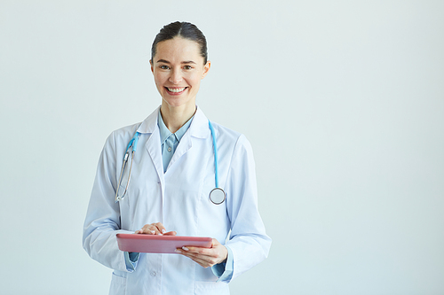 Waist up portrait of smiling female doctor holding clipboard and  while standing against white wall in clinic, copy space