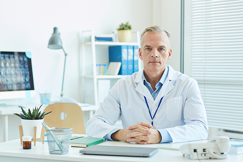 Portrait of mature male doctor wearing lab coat and  while sitting at desk in office of modern clinic, copy space