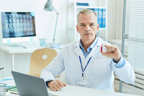 Portrait of mature male doctor holding blood test sample and  while sitting at desk in office of modern clinic, copy space