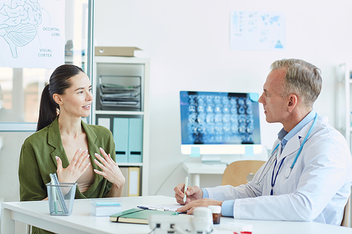 Portrait of emotional young woman talking to mature doctor while sitting at desk during consultation in modern clinic, copy space