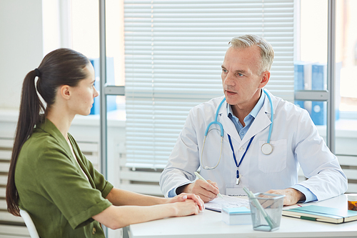 Portrait of mature male doctor talking to young female patient while sitting at desk during consultation in modern clinic, copy space