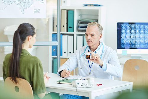 Portrait of mature male doctor holding pill bottle while sitting at desk and consulting female patient in modern clinic, copy space
