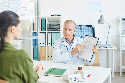 Portrait of mature male doctor pointing at chart while consulting female patient in modern clinic, copy space