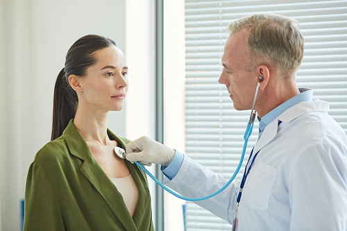 Side view portrait of mature doctor listening to breath and heartbeat of young woman via stethoscope while examining patient in clinic