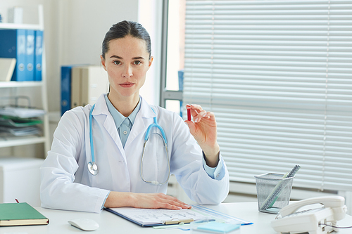 Portrait of professional female doctor holding blood test sample and  while sitting at desk in office of modern clinic, copy space