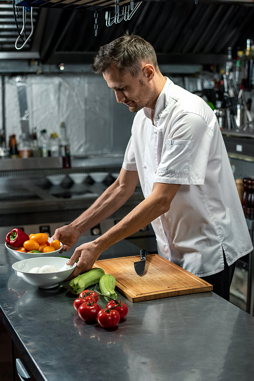 young professional chef of modern restaurant in white uniform standing by table in the kitchen and preparing ingredients for  garnish