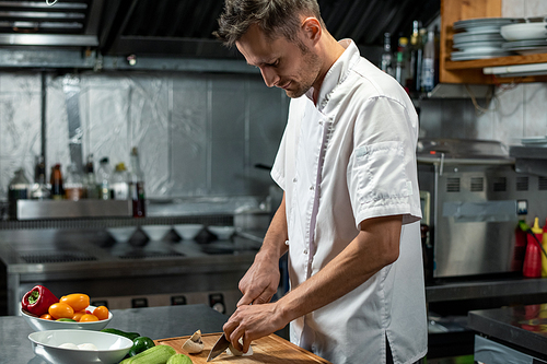 young professional chef of modern restaurant in uniform standing by large table in the kitchen while cutting mushrooms and other s