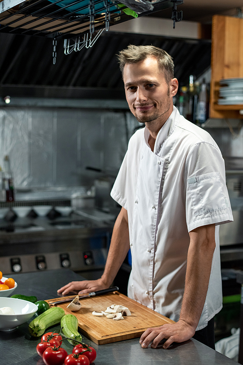 young successful professional chef of modern restaurant in white uniform standing by table in the kitchen while cooking  coarse