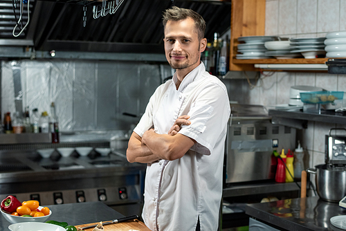 Young successful chef of modern restaurant in white uniform crossing arms by chest while standing by table in the kitchen before cooking salad