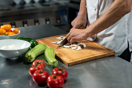 Hands of young male chef of modern restaurant in white uniform standing by large table in the kitchen and cutting mushrooms on wooden board