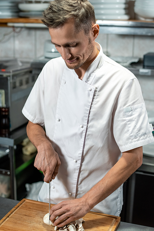 young male chef of modern restaurant in white uniform chopping mushrooms for  garnish while standing by table in the kitchen