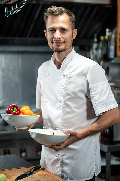 young successful professional chef of modern restaurant in white uniform holding two ceramic bowls with fresh capsicum for  course