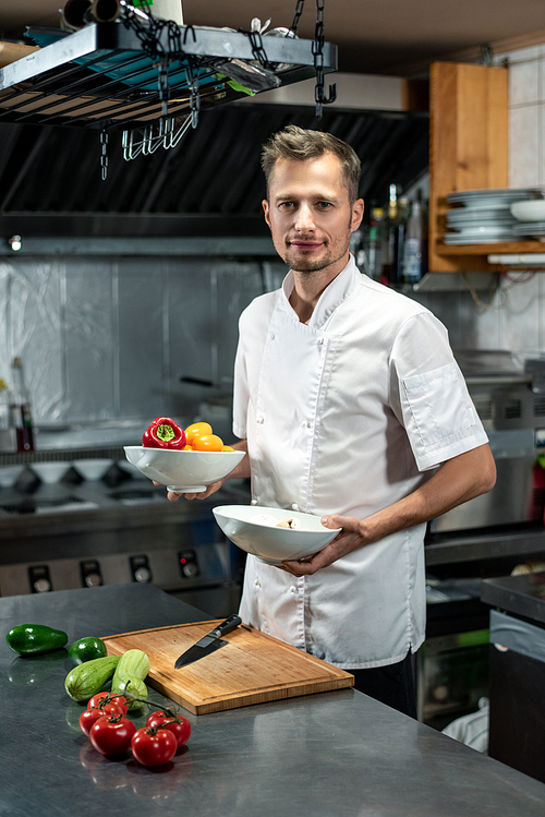 happy young professional chef standing by table and holding two ceramic bowls with fresh capsicum for  salad or other course