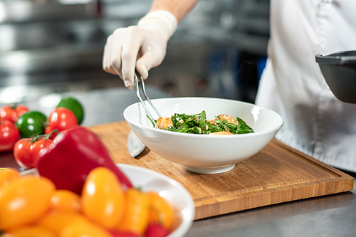 Gloved hand of young chef with fork putting cooked meal into white ceramic bowl standing on wooden board surrounded by fresh vegs