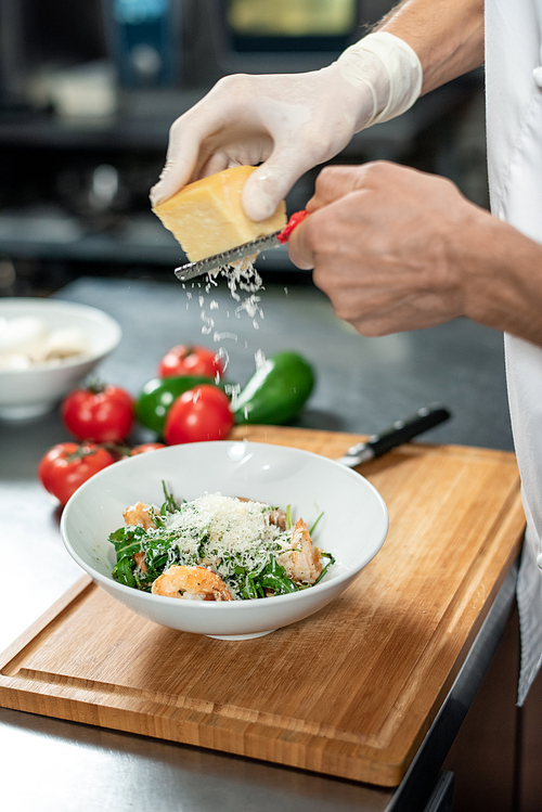 Hands of chef in uniform grating fresh cheese into ceramic bowl containing salad consisting of ruccola, boiled shrimps and other ingredients