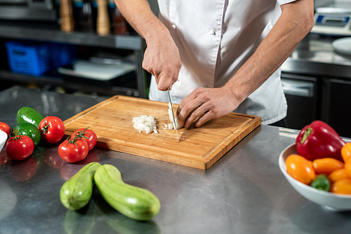 Hands of young male chef in uniform cutting fresh onions on wooden board surrounded by tomatoes and zucchini while cooking lunch