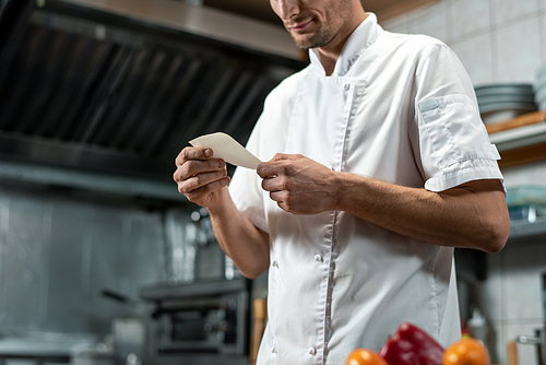 young chef in white uniform looking through list of ingredients for  salad or other dish while cooking food in the kitchen