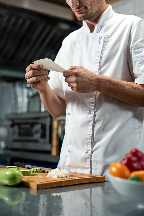 young chef in uniform looking through list of ingredients for  salad while holding paper over chopped onion on wooden board