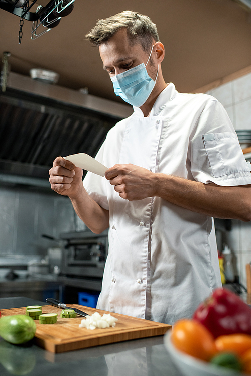 Young chef in protective mask standing by table with chopped onion and zucchini on board while looking through list of ingredients for salad