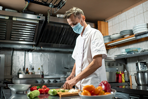 professional young chef in uniform and protective mask chopping fresh zucchini and other s while standing by large table