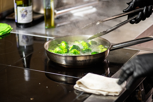 Close-up of unrecognizable chef in black gloves using tongs while frying broccoli on cooking pan