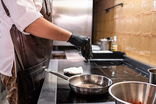 Close-up of unrecognizable chef in apron and gloves standing at stove and adding salt in cooking pan