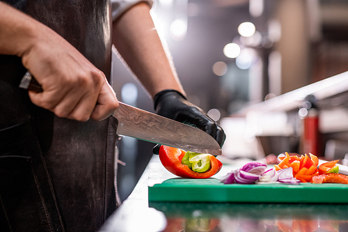 Close-up of unrecognizable chef in apron cutting bell pepper with kitchen knife while making refreshing summer salad