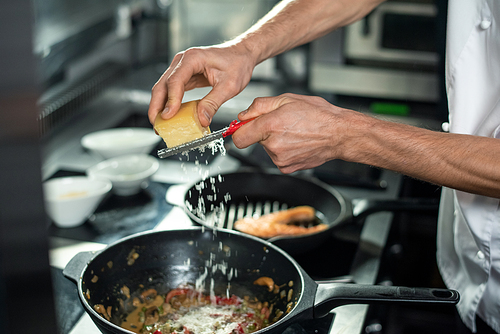 hands of young professional chef in white uniform grating cheese into frying pan with chopped s while cooking in the kitchen