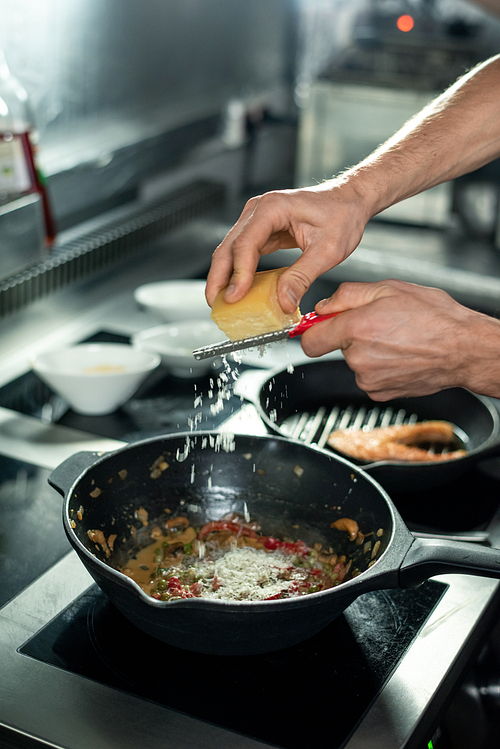 hands of young professional chef grating cheese into frying pan with chopped s while cooking stew for client in the kitchen