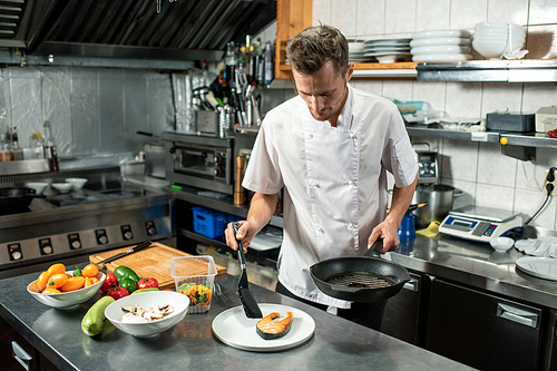 Contemporary male chef putting piece of fried salmon on plate while standing by large kitchen table and preparing meal for client
