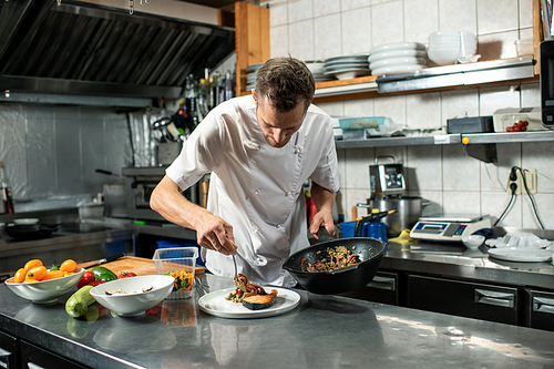 young male chef in uniform holding frying pan with  stew while bending over table with plate and putting food on piece of fried salmon
