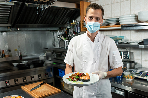 young male chef in white uniform and protective mask holding plate with fried salmon and s while standing in front of camera