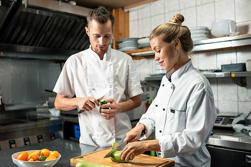 Young confident professional chef with knife peeling avocado and consulting his female trainee cutting fresh zucchini on wooden board
