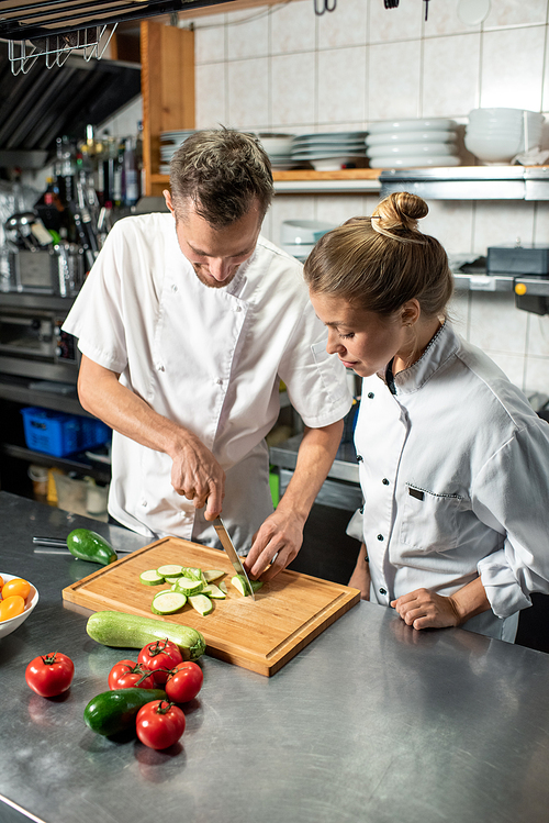 Young professional male chef showing his female trainee how to cook fresh zucchini while both standing by table in the kitchen of restaurant