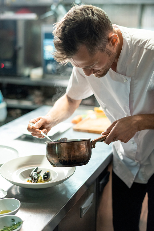 Young male chef in uniform bending by kitchen table and putting boiled mussels on plate while taking them out of metallic pan before serving