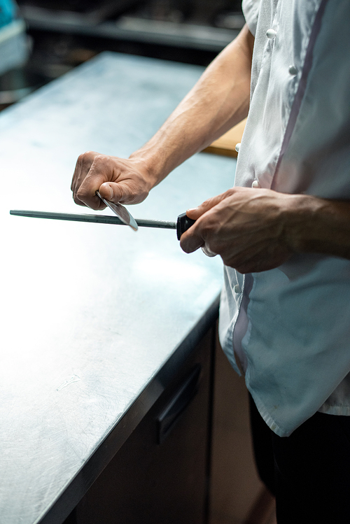 Hands of young contemporary chef of restaurant in white uniform sharpening knife over kitchen table before preparing meal