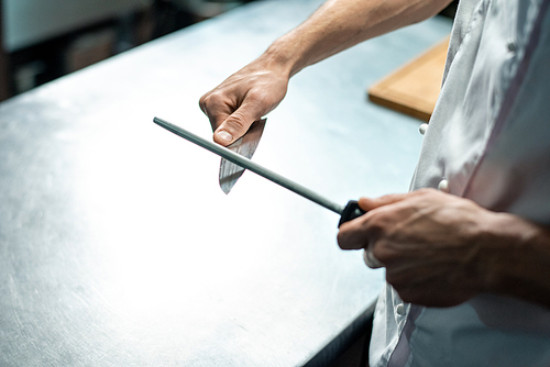 Hands of young male chef of modern restaurant in white uniform sharpening knife over kitchen table before chopping raw ingredients