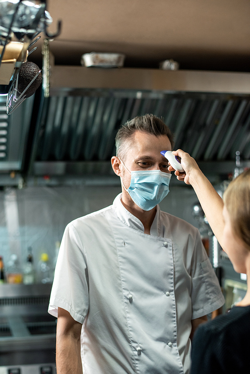 Contemporary chef in uniform and protective mask looking at woman with thermometer measuring his body temperature before work in the kitchen