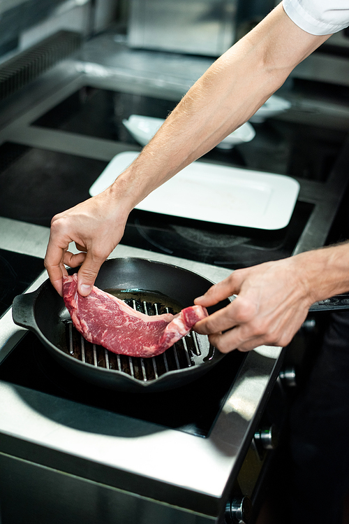 Hands of young chef putting piece of meat on hot grill frying pan with vegetable oil while standing by electric stove and cooking beef