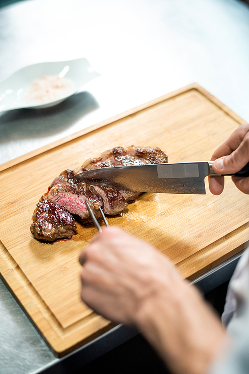 Hands of chef with sharp knife and fork cutting roasted beef steak on wooden board before putting it on plate with garnish and serving