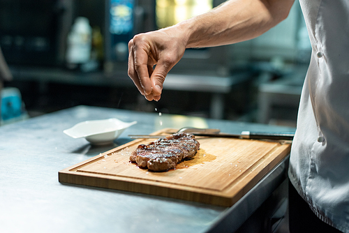 Hand of chef sprinkling spices on roasted beef steak on wooden board while standing by table and preparing meal for client of restaurant