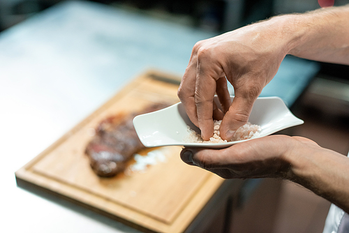 Hands of chef taking marine salt to add it to roasted beef steak on wooden board while standing by table and preparing meal for client