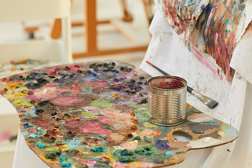 Background image of colorful artists palette covered with paint blots resting by easel in art studio lit by sunlight, copy space
