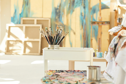 Background image of set of paintbrushes and tools resting by easel in empty art studio lit by sunlight, copy space