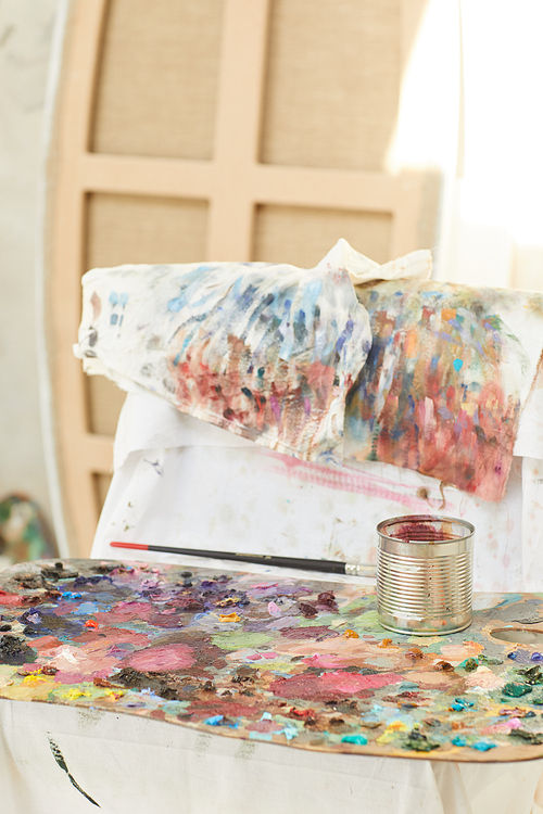Vertical background image of colorful artists palette covered with paint blots resting by easel in art studio lit by sunlight, copy space