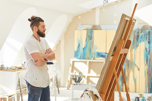 Side view portrait of contemporary male artist looking at painting while standing by easel in sunlit art studio, copy space